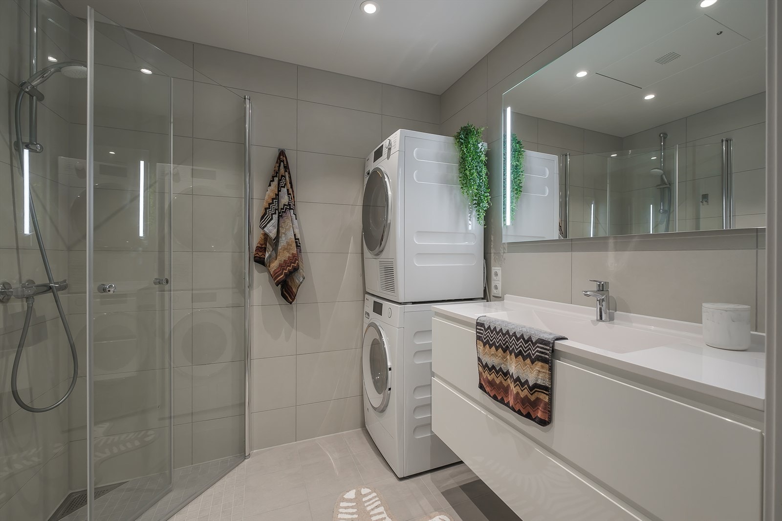 Lyst og delikat baderom utstyrt med stor servantinnredning, speil med lys, dusjhjørne og vegghengt toalett, samt opplegg for vaskemaskin. Galleribilde