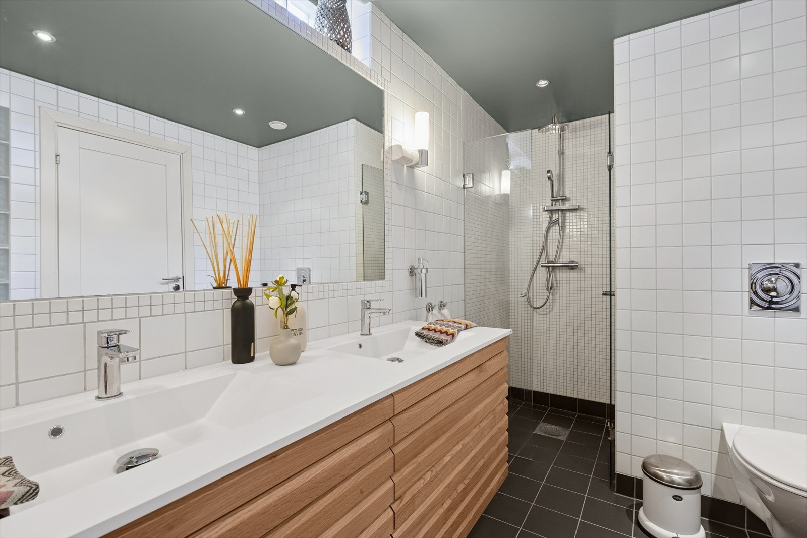 Pent, flislagt baderom med innredning med to nedfelte servanter, veggmontert toalett og dusjvegger/hjørne. Galleribilde