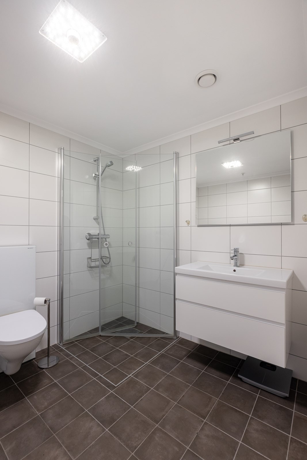 Innredning med dype skuffer og heldekkende servant, samt speil med overlys. Badet er også innredet med dusjnisje med plassbesparende innfellbare glassdører og veggmontert toalett. Galleribilde