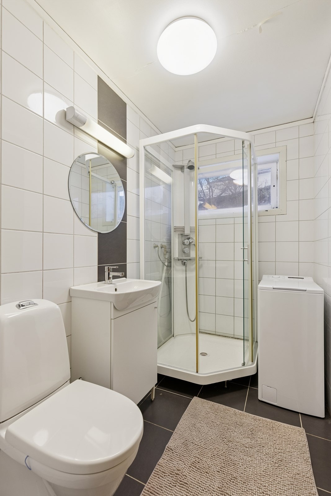 Baderom i hybeldel med opplegg for vaskemaskin, utstyrt med gulvstående klosett, dusjkabinett og servantinnredning. Speil og belysning på vegg. Galleribilde