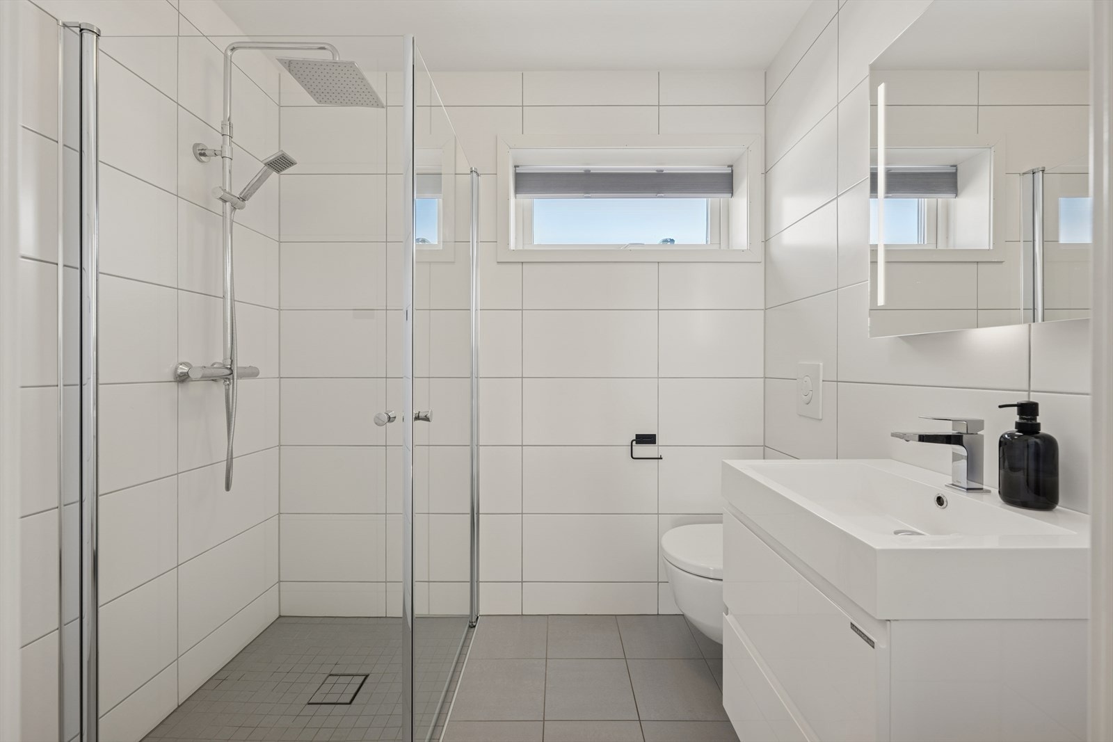 Rommet er innredet med et stilrent dusjhjørne med glassvegger, servant på praktisk skuffeinnredning, speil med integrert belysning og et moderne vegghengt toalett. Galleribilde