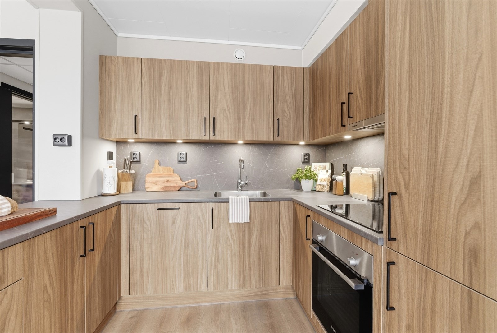 Kjøkkenet er særdeles delikat, med slette fronter i lun trefinér, ton-i-ton benkeplate og mørke detaljer. Innredningen har integrerte hvitevarer som medfølger salget. Galleribilde