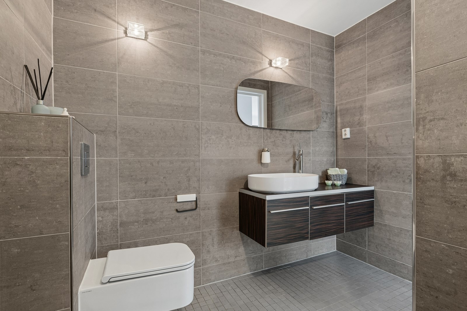 Lekkert baderom bestående av servant med underskap, speil med lys over, frittstående badekar og veggmontert toalett med innebygget sisterne Galleribilde