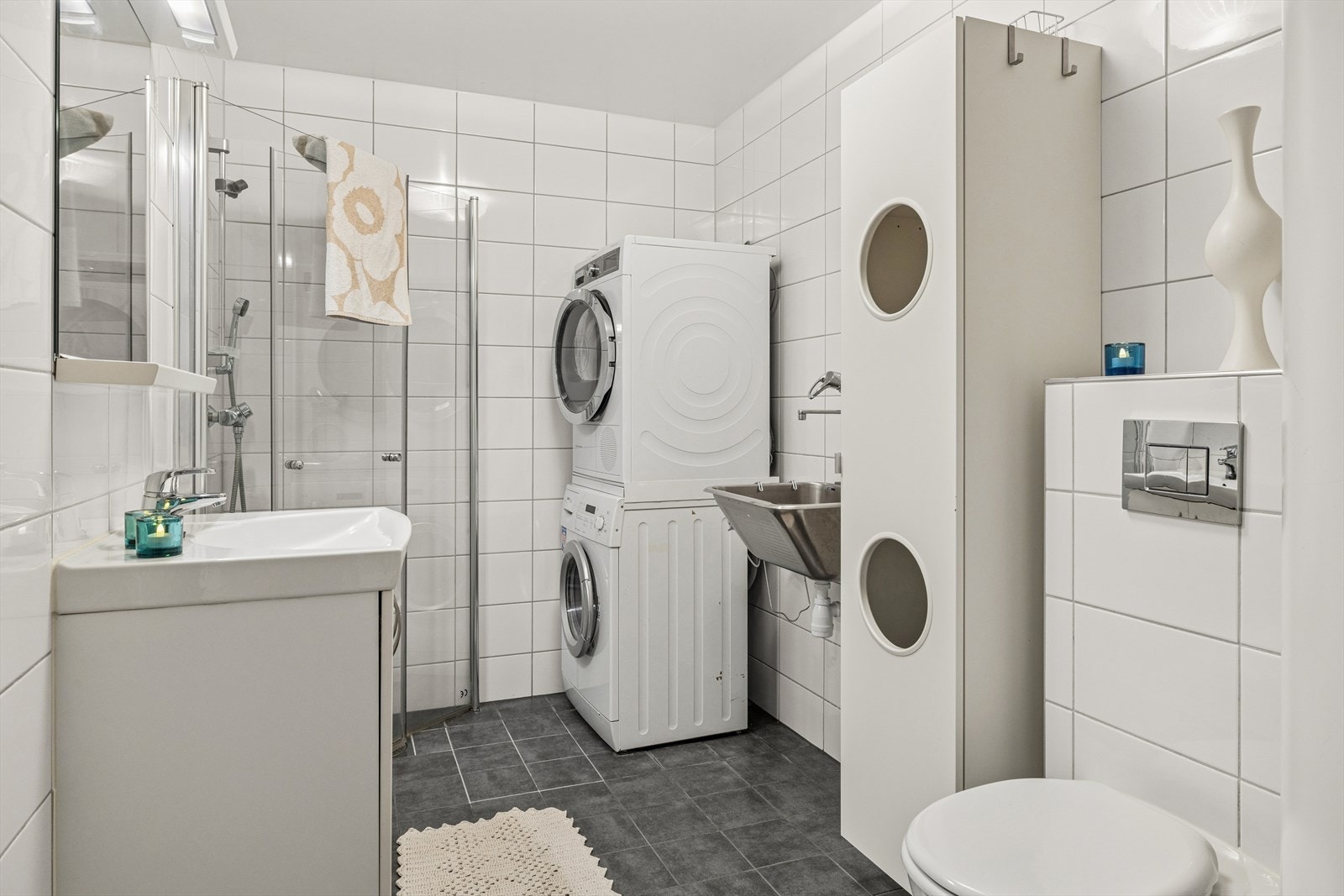 Flislagt bad med innredning bestående av servant, speil og veggmontert toalett med innebygget sisterne. Opplegg for vaskemaskin Galleribilde