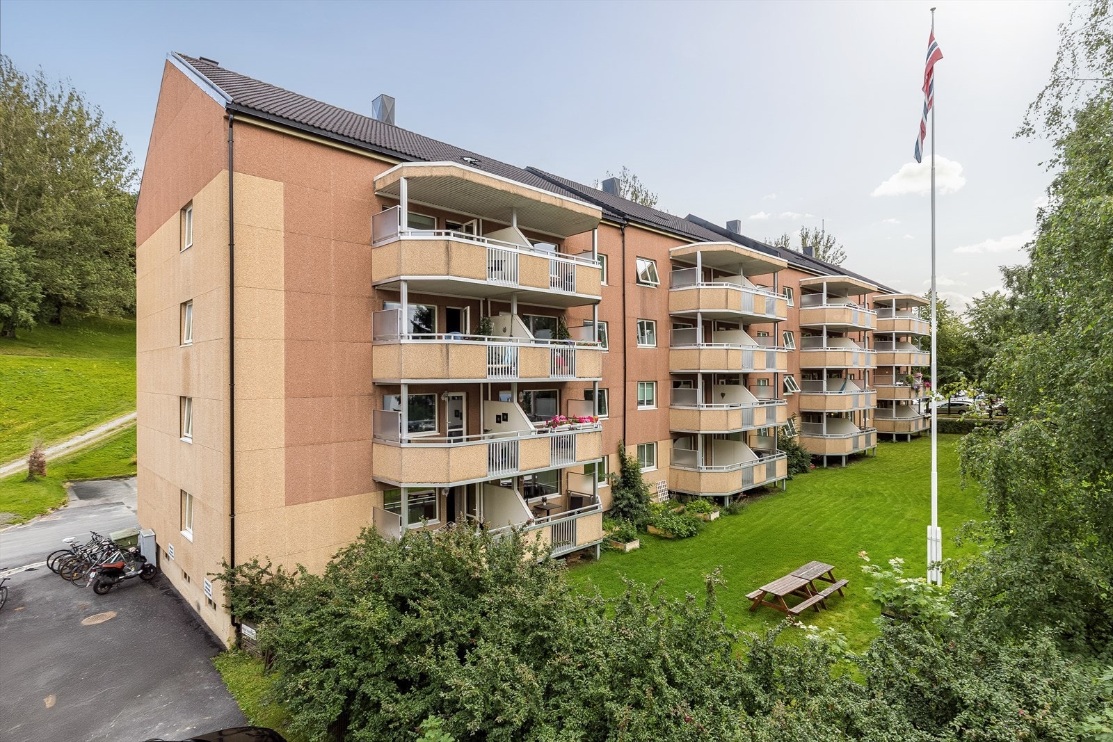 Sommerbilde av fasade. Vinduer og dører i bygget ble skiftet i 2014. Området har gode kollektive transportmuligheter, og nærmeste busstopp ligger ca. 250 meter fra leiligheten. Galleribilde