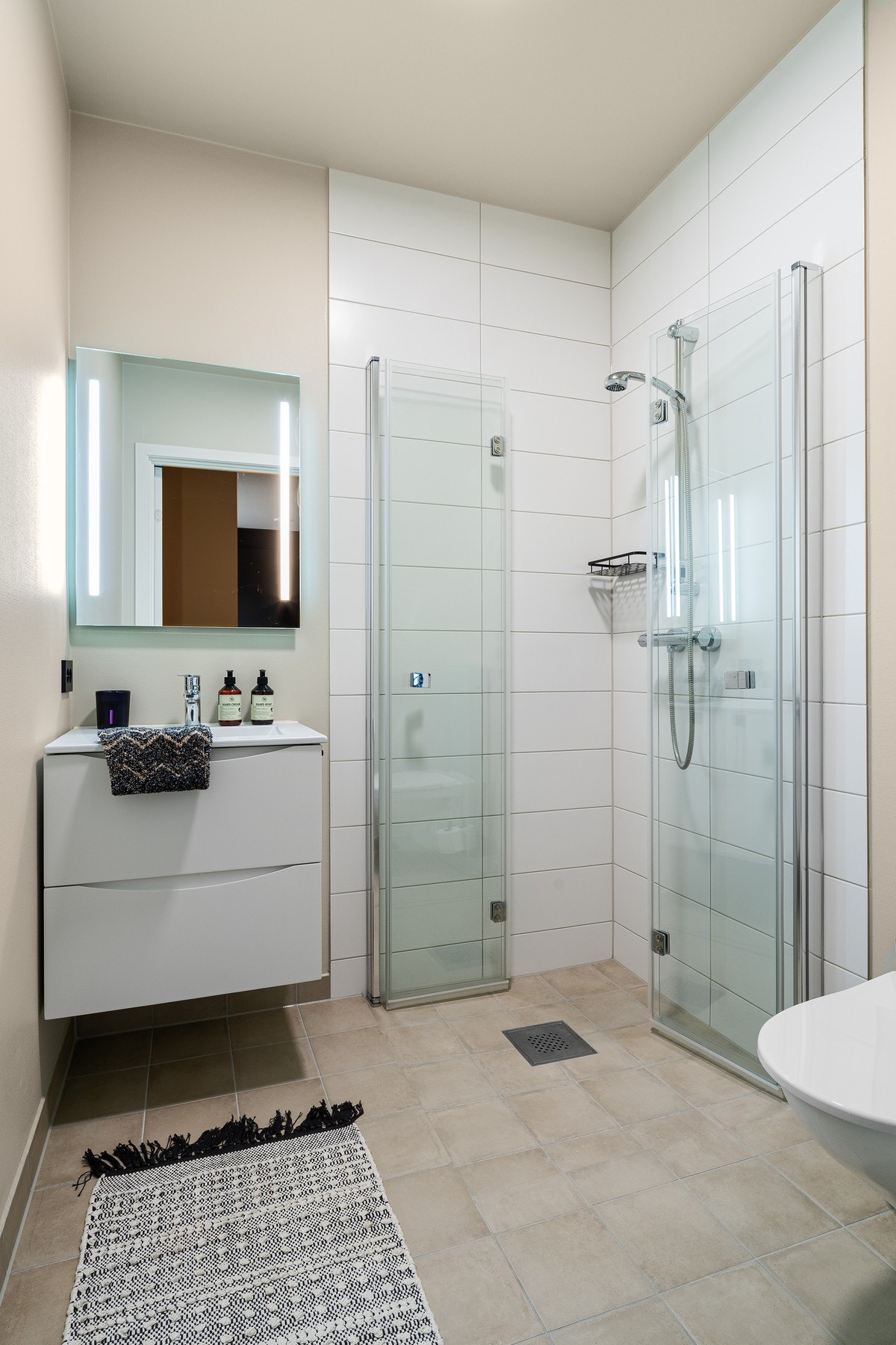 Badet i første etasje har fliser på gulv og vegger i dusjsonen. Vegger ellers med malt strietapet. Badet er innredet med servant på skuffeseksjon, speil med belysning. Videre er badet utstyrt med dusjdører og vegghengt toalett. Galleribilde