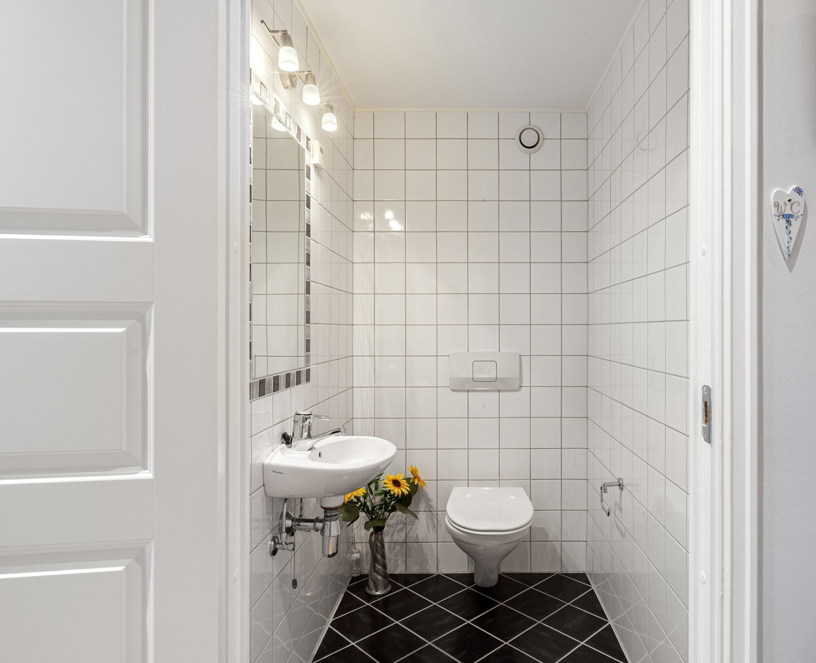 Vegg i vegg med badet ligger et toalettrom. Det er fliser på gulv og vegger, samt malte flater i taket. Rommet har vannklosett, servant, speil og lys. Galleribilde