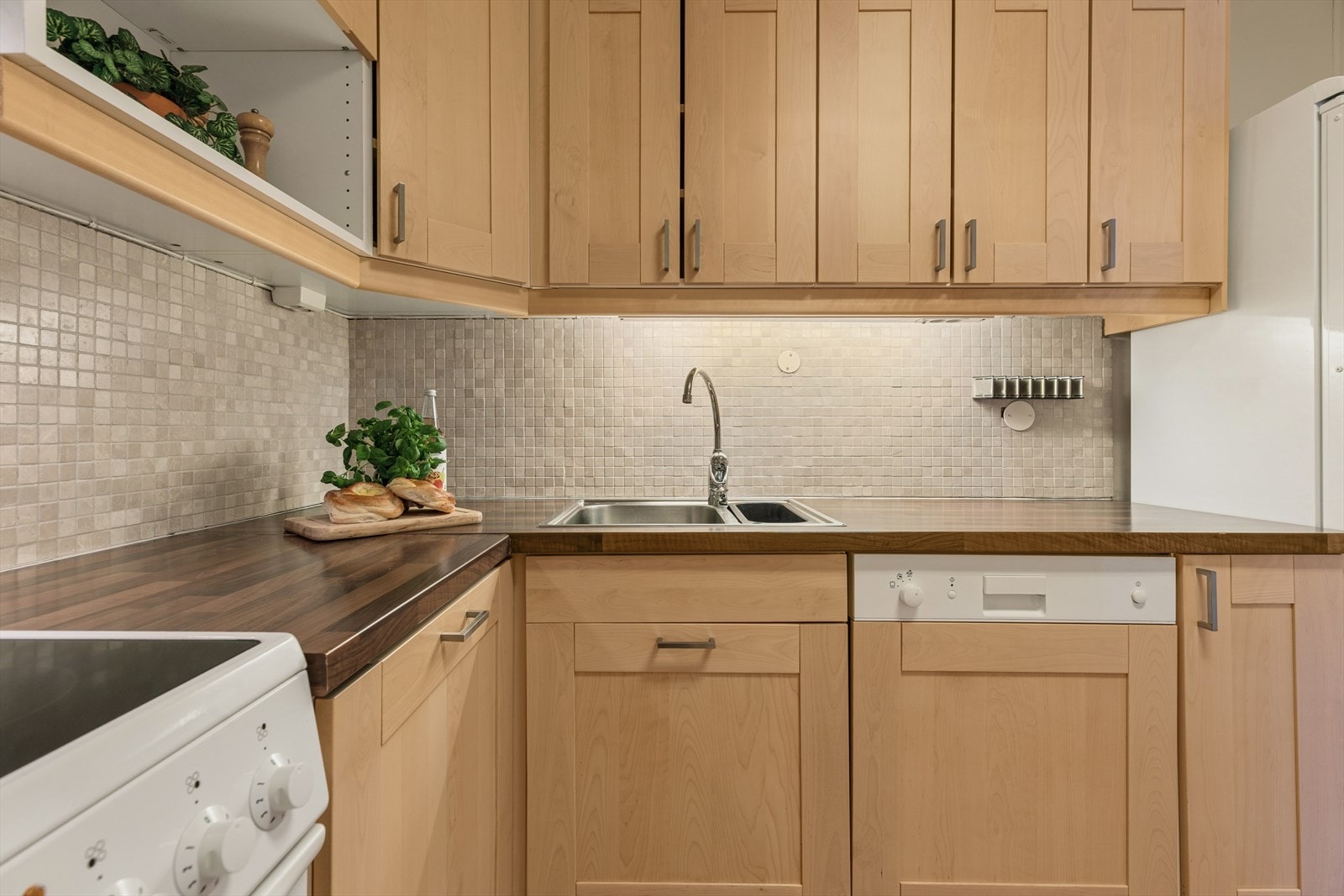 Benkeplaten har nedfelt stålkum med to basseng og ettgreps kjøkkenkran. Mellom benk og overskap er det fliser som gir et slitesterkt og lettstelt arbeidsområde Galleribilde