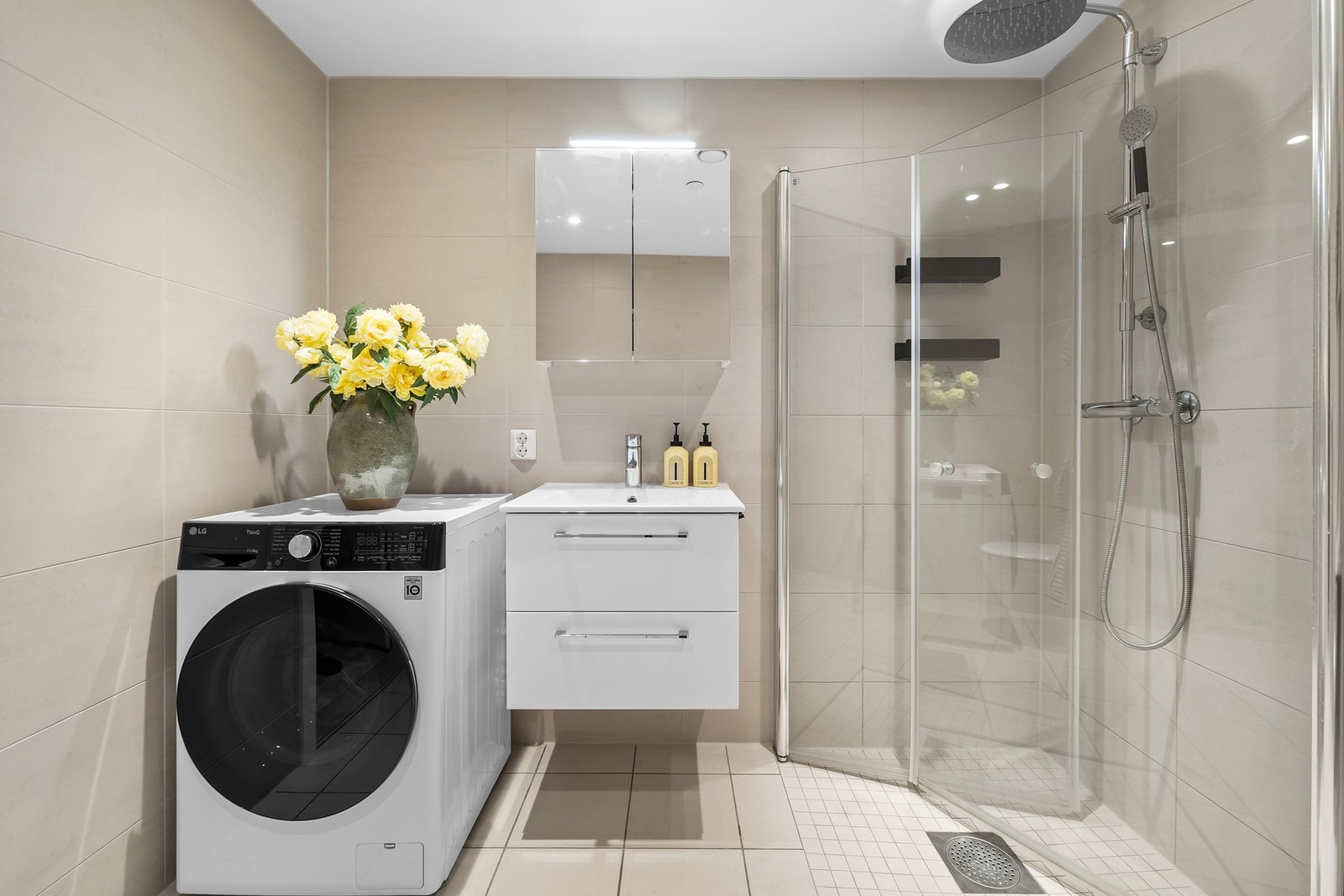 Flislagt bad med moderne uttrykk, utstyrt med dusjløsning, servant med innredning og opplegg for vaskemaskin. Galleribilde