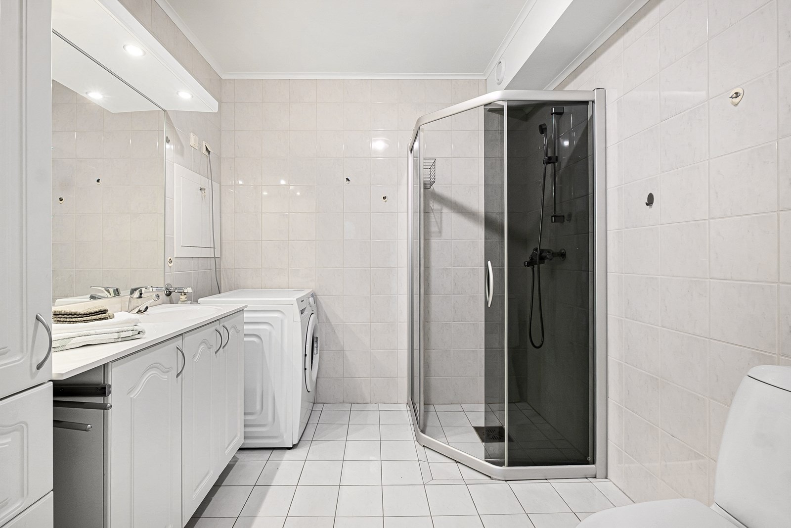 Lyst og pent flislagt bad utstyrt med dusjhjørne, gulvstående toalett og opplegg for vaskemaskin. Galleribilde