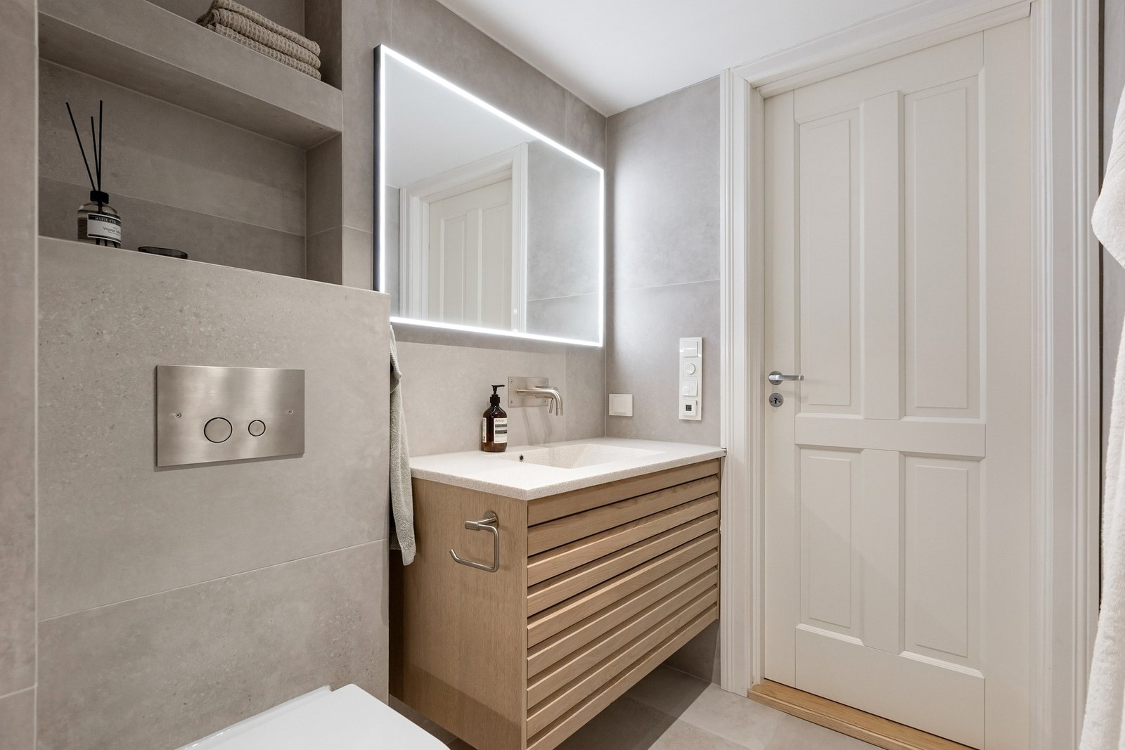 Videre inneholder baderommet servantskap med glatte fronter, heldekkende servant, speil med lysarmatur på vegg, veggmontert klosett, samt dusjhjørne med to veggmonterte armatur, hjelpesluk og termostatstyrt blandebatteri. Galleribilde