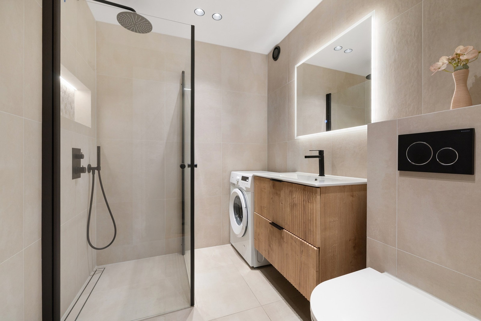 Delikat, flislagt baderom med gulvvarme og downlights i himling. Dusjhjørne med innfellbare glassdører og integrert dusjsett med rainshower i sort utførelse og innebygd nisje med plass til dusjsåpe Galleribilde