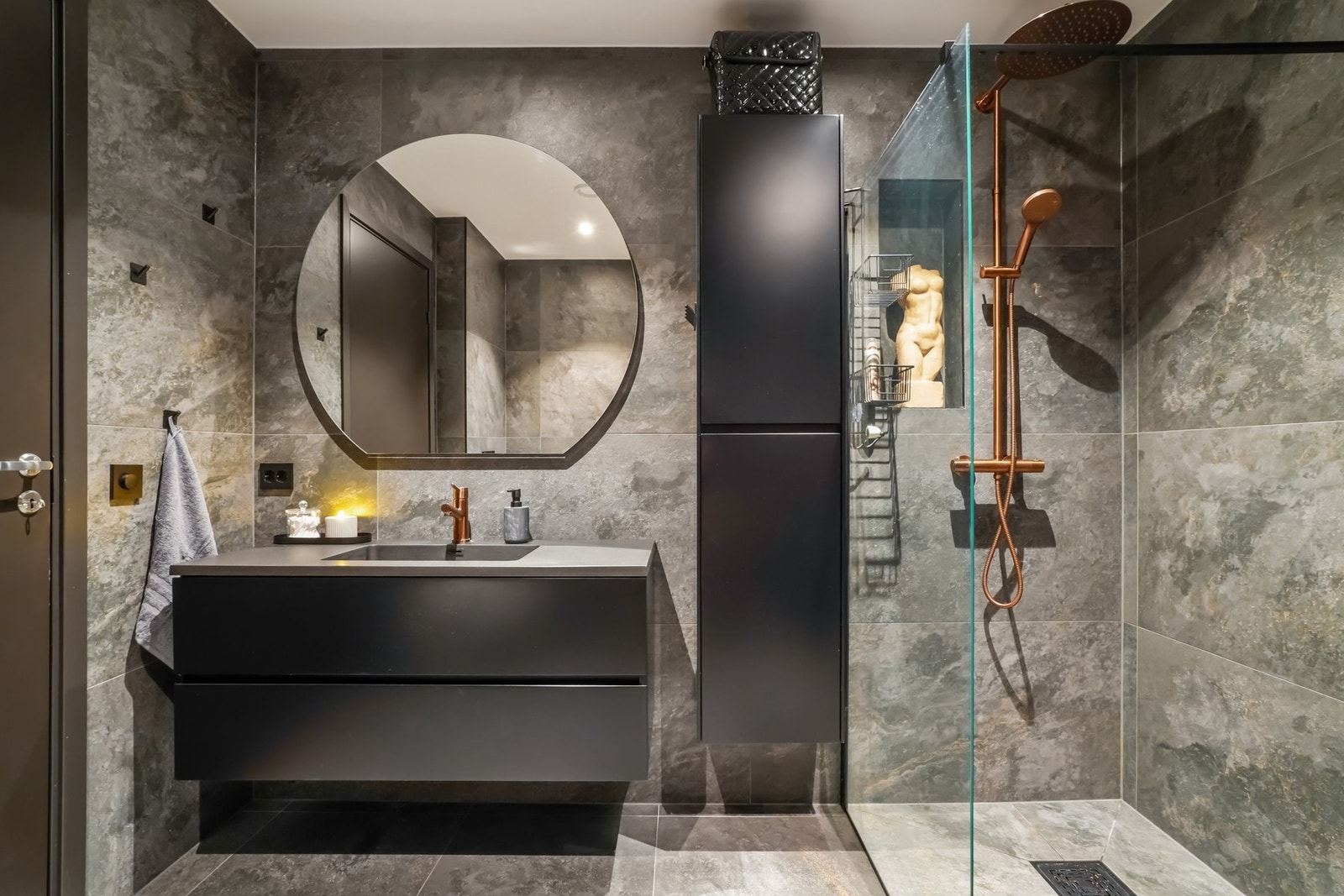 Servantinnredning med to mørke skuffer, mørk helstøpt servantplate, rundt speil med belysning bak, høyskap og dusjhjørne med glassvegg. Galleribilde