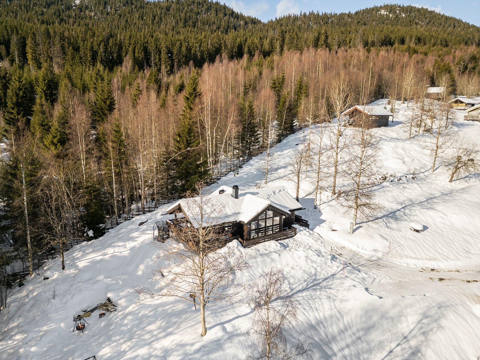 Fra luften fremheves den harmoniske plasseringen i terrenget, med åsrygger og skogsområder som omkranser hytta og skaper en følelse av privatliv og ro. Hytta skiller seg tydelig ut med sin moderne arkitektur, store glassflater og brunbeisede fasade. Galleribilde