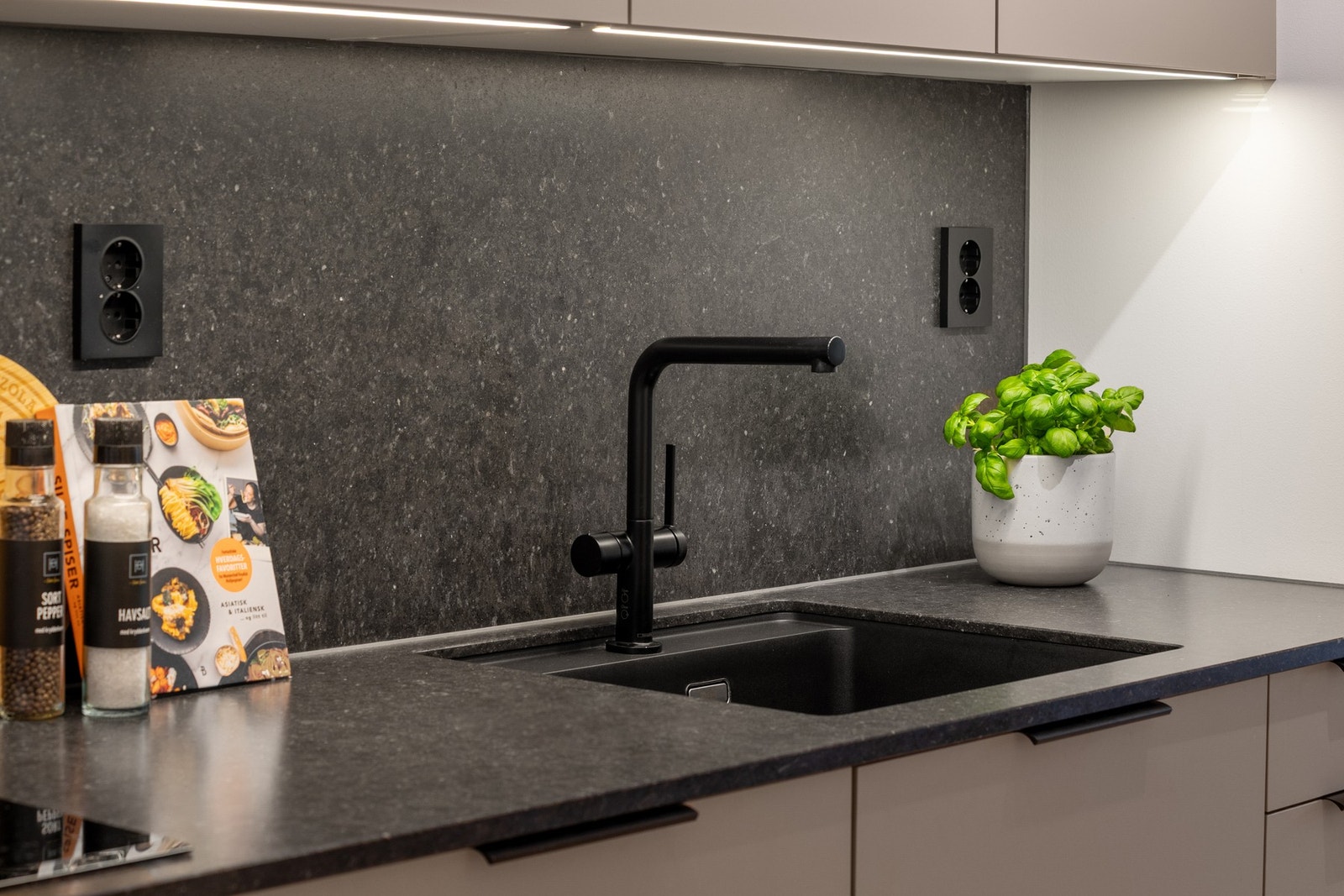 Detaljbilde fra kjøkkenet. Kjøkkenet er oppgradert med blant annet benkeplaten, underlimt vask, kjøkkenarmatur, stikkontakter, lyslist under overskap, håndtak og fronter. Galleribilde