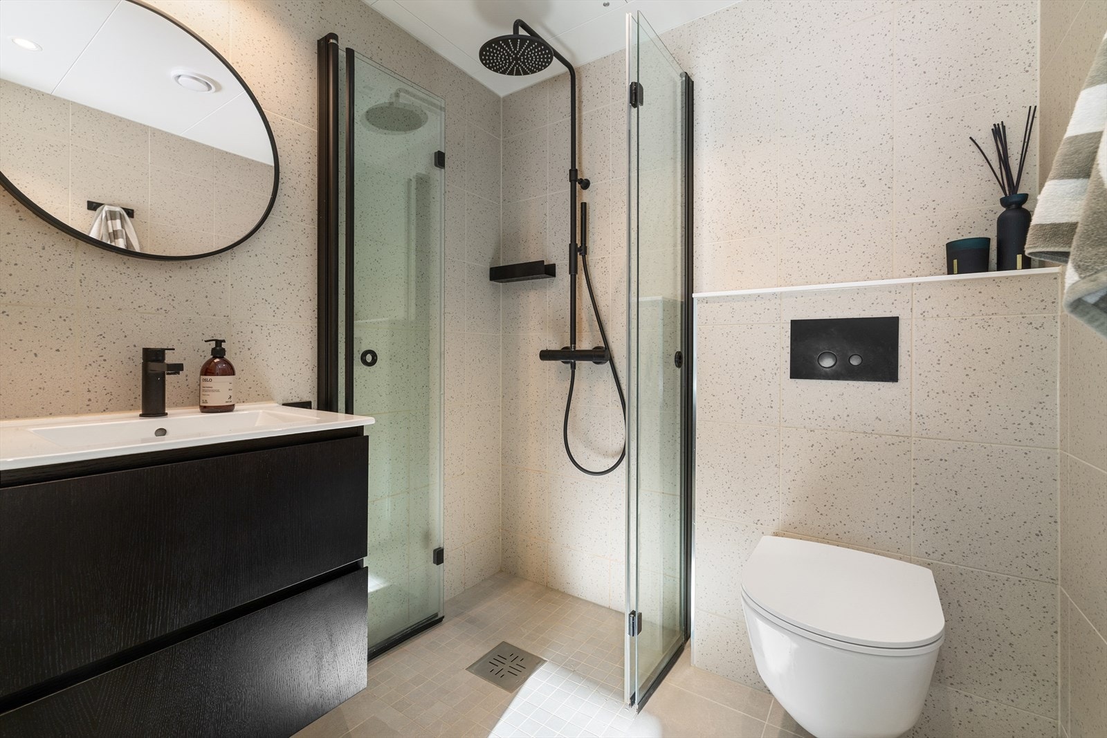Lekkert flislagt bad med lyse fliser og sorte detaljer på innredning. Det er gjort tilvalg på regnfalldusj og trykkplate til toalett. Oppgradert speilbelysning. Galleribilde