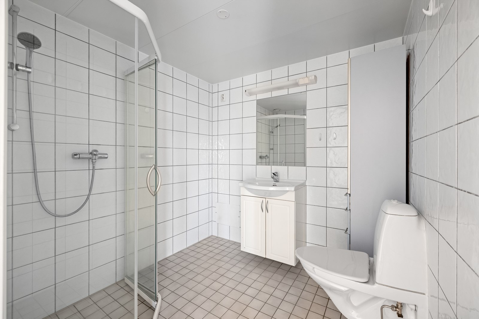 Bad med flislagte vegger og gulv, dusjhjørne med glassdører, servant med underskap og opplegg for vaskemaskin. Galleribilde
