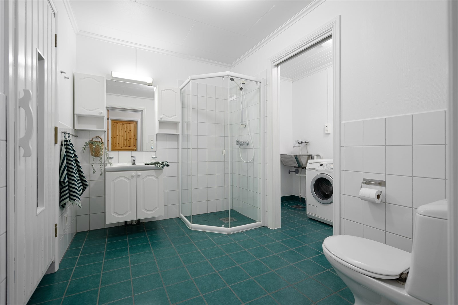 Flislagt bad i 1. etasje med elektrisk varme i gulv, dusjhjørne, toalett, servant med benkeskap og tilgang til vaskerom. Galleribilde