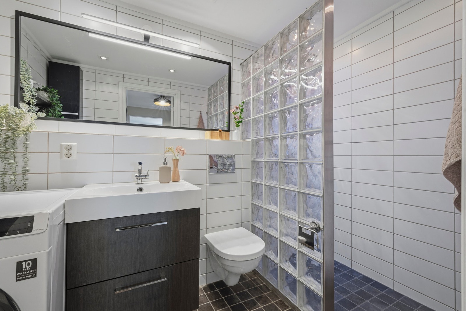 Rommet har en vegg av glassbyggerstein som skjerming mot dusjnisje og et stort speil med belysning over servantseksjon Galleribilde