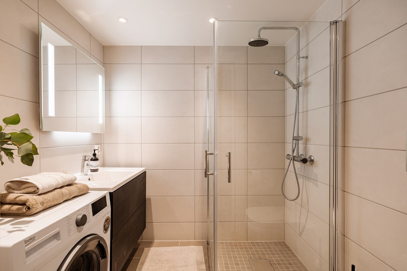 Moderne, flislagt bad med en slett innredning med heldekkende servant, speil med integrert lys opplegg for vaskemaskin. Galleribilde