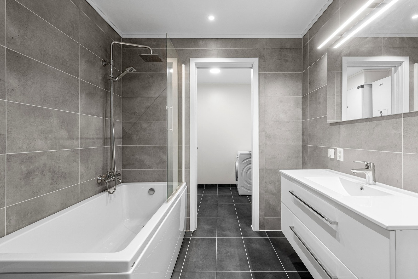 Romslig og moderne bad med god funksjonalitet og gjennomgang til vaskerom. Her finner man baderomsinnredning med servant, vegghengt toalett, badekar samt opplegg for vaskemaskin og tørketrommel. Galleribilde