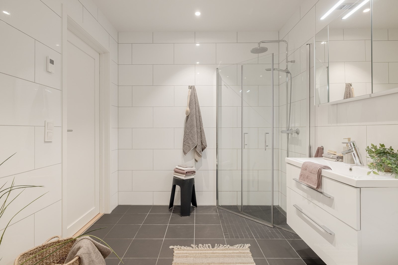 Badet er utstyrt med dusjnisje med innfellbare dører og vegghengt dusjbatteri med regnfall. Baderomsinnredning med skuffer og heldekkende servant. I overkant av servanten er det speilskap med lyslist. Galleribilde