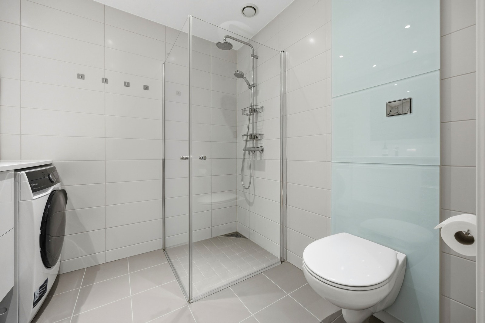 Separat dusjsone med regnfallsdusj og innfellbare dusjvegger i glass. Det er opplegg og plass for vaskemaskin og tørketrommel under praktisk benkeinnredning. Galleribilde