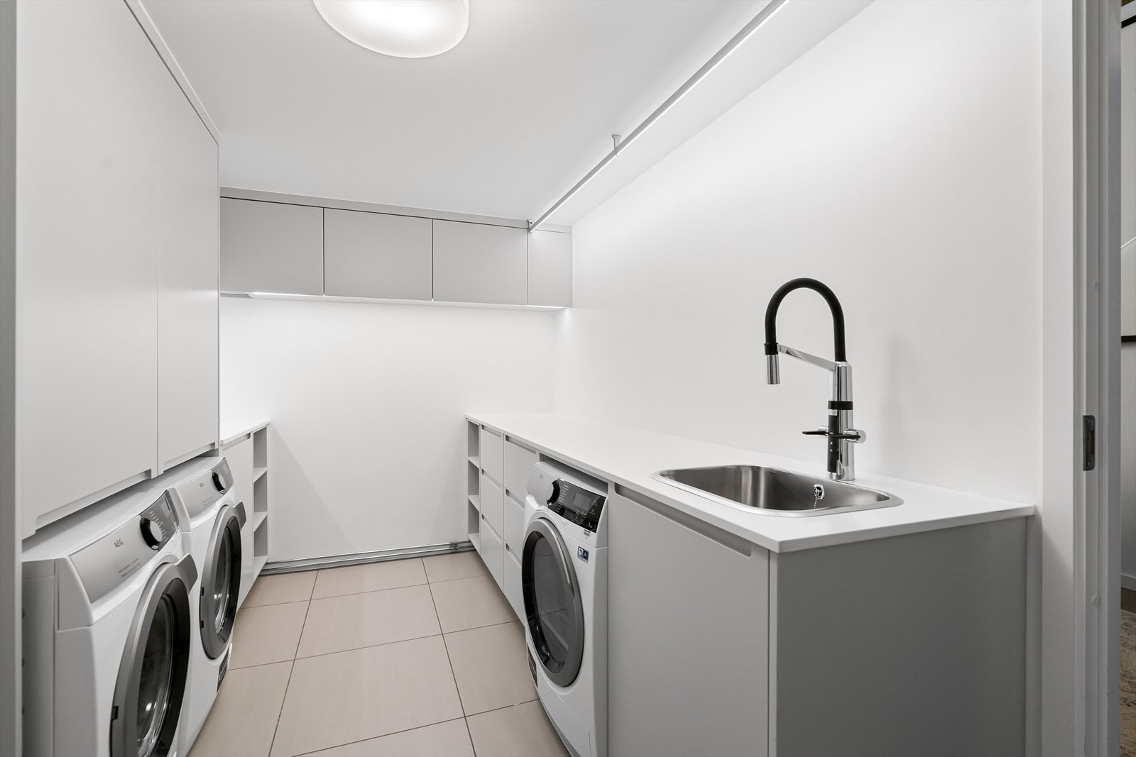 Svært praktisk vaskerom med innredning med godt med skap- og benkeplass, fliser på gulvet og plass til flere vaskemaskiner og tørketrommel. Galleribilde