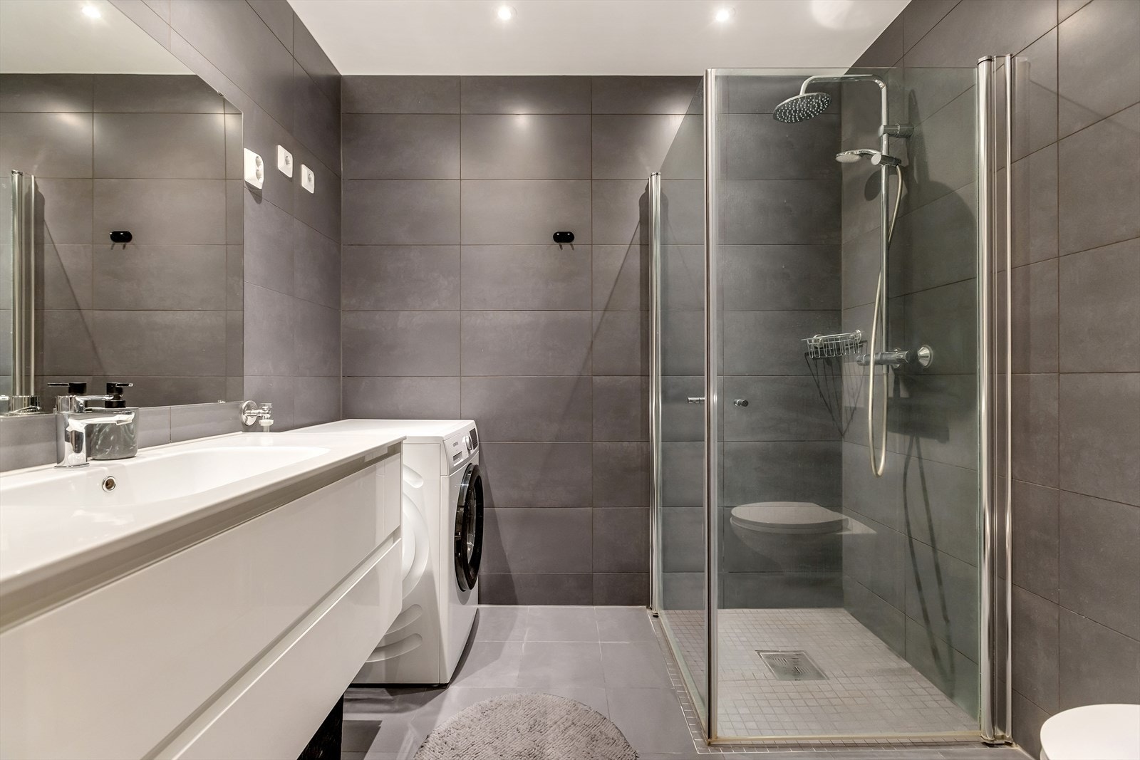 Badet fremstår moderne og stilrent, med store vegg- og gulvfliser i grå nyanser. Dusjsonen har mosaikkfliser, regnfallsdusj og praktiske innfellbare glassdører. Galleribilde