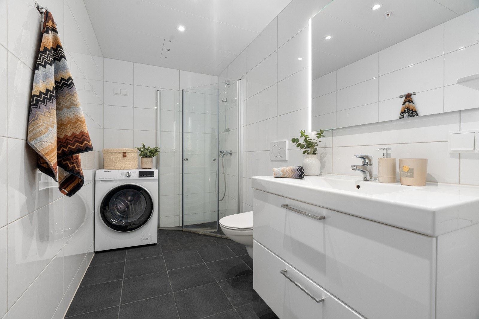 Lekkert flislagt bad - utstyrt med praktisk vegghengt wc, stort dusjhjørne og opplegg for vaskemaskin. Baderomsinnredning med god oppbevaringsplass.