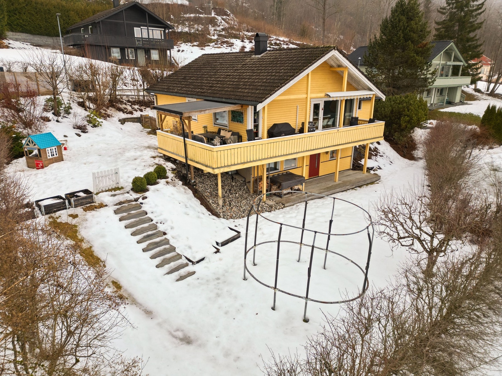 Fin og skjermet hage over to nivåer med trapp mellom nivåene. Under snøen på bildene er hagen pent opparbeidet med grøntarealer og gruset innkjøring. Det er stor veranda og terrasse med god plass til grill og overbygget pergola på verandaen. Galleribilde