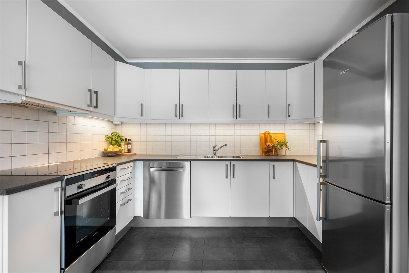 Hvit, glatt innredning fra HTH med laminat benkeplate, oppvaskmaskin, stekeovn m/flatetopp, oppvaskkkum i stål og frittstående kjøl/fry. Kjøkkenet har godt med skap- og benkeplass Galleribilde