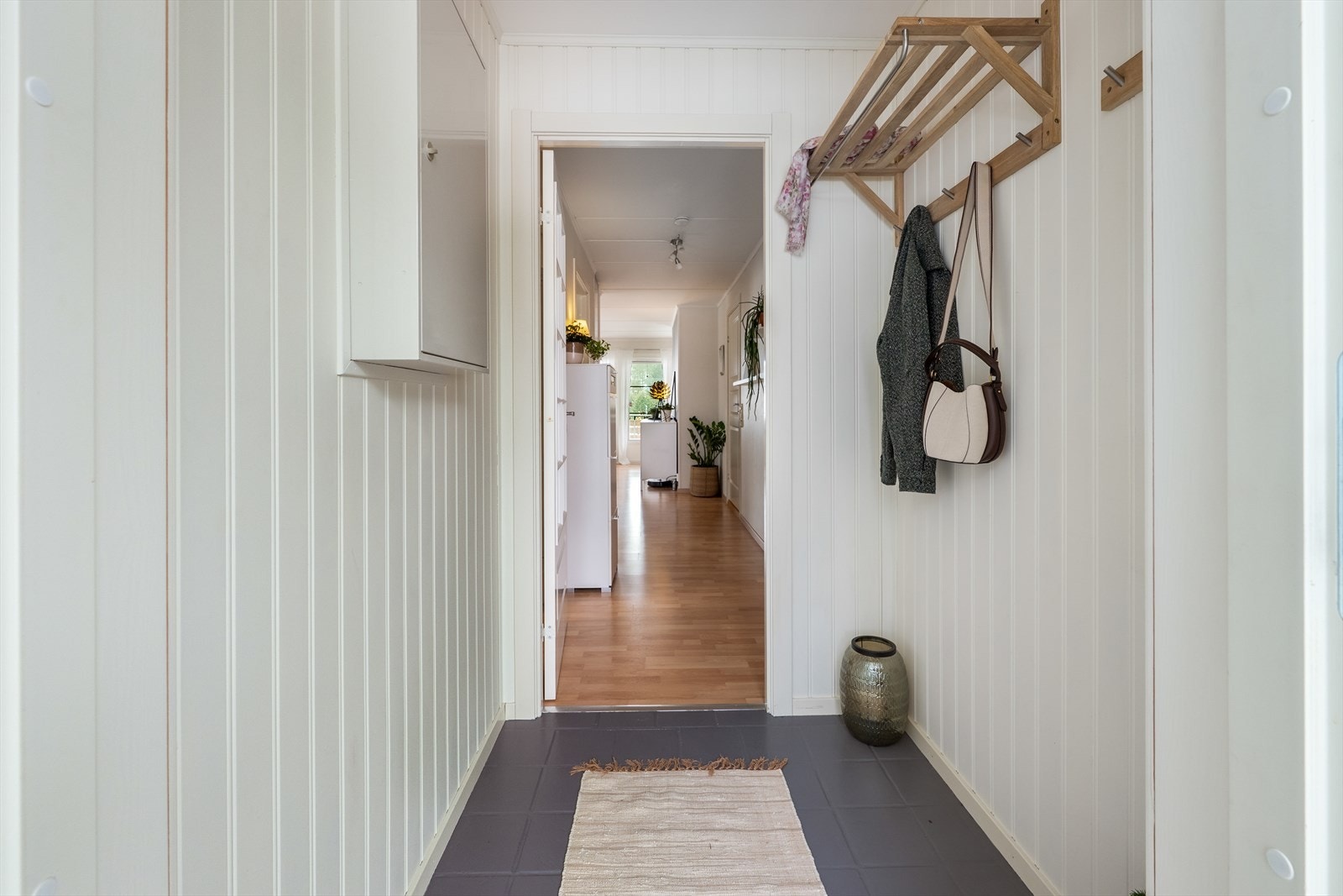 Entreen er pent innredet med fliser på gulv, lysmalt panel på vegger og fin plass til knaggrekke og skoskap Galleribilde