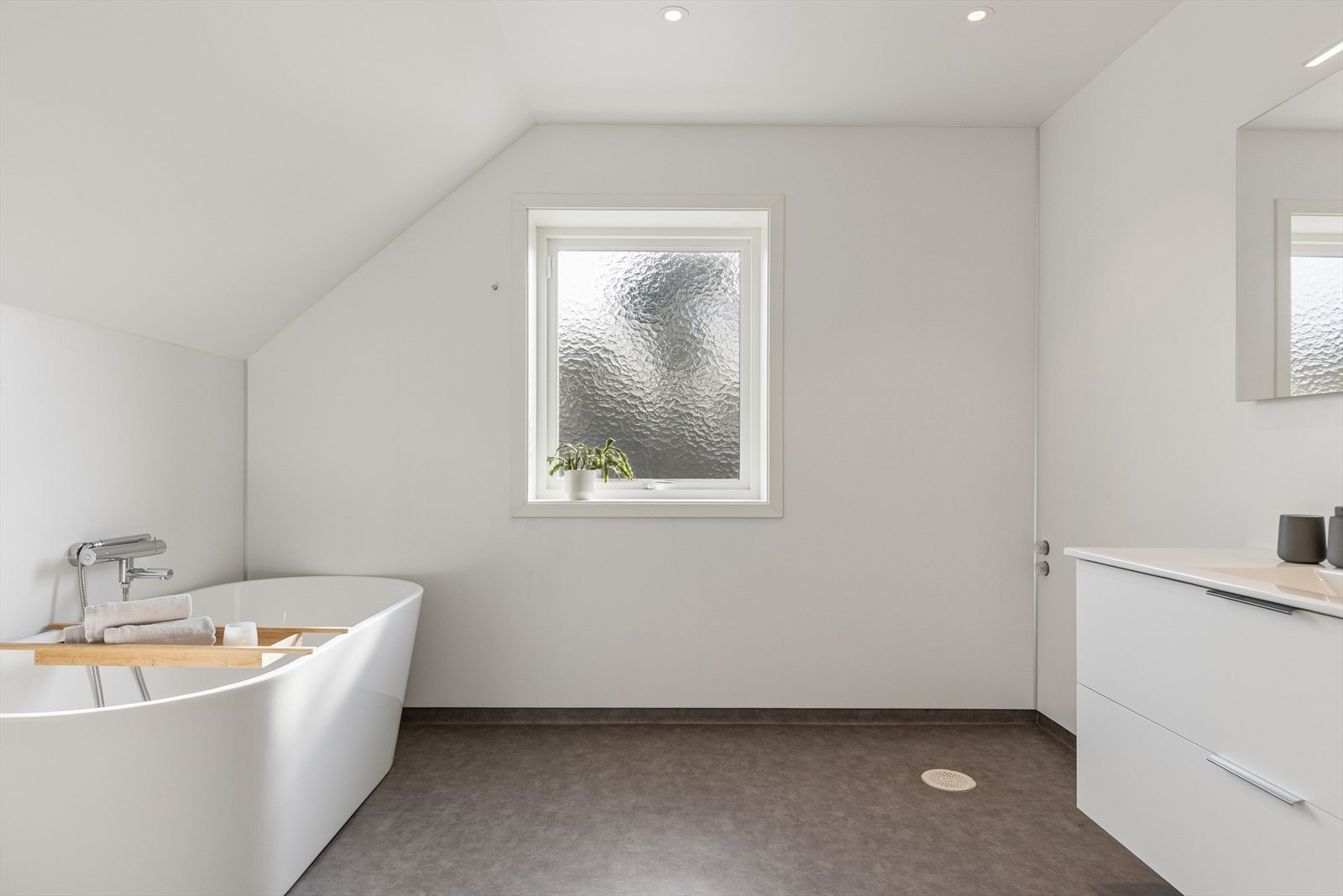 Stort, innbydende bad med gulvbelegg på gulv og våtromsplater på vegger. Rommet har baderomsinnredning, badekar, fordelerskap for vannrør og veggmontert toalett. Galleribilde