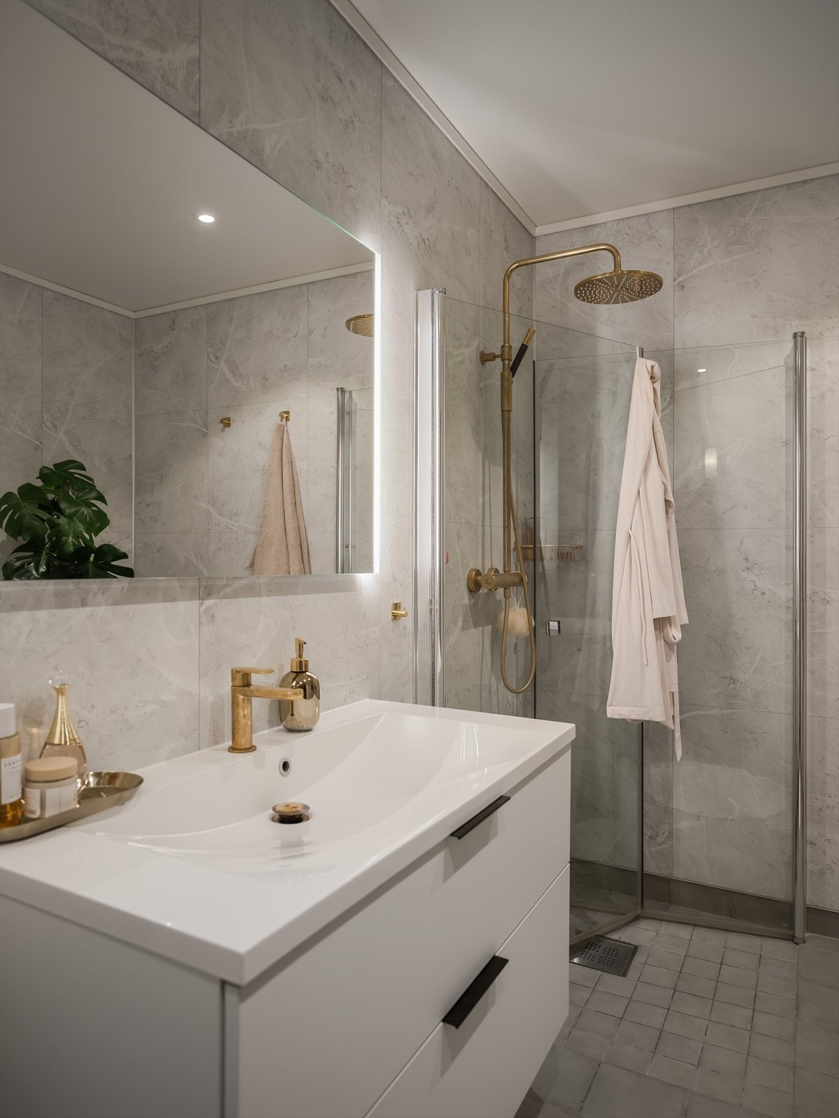 Badet er innredet med hvit servantinnredning med skuffer til oppbevaring, toalett, dusjhjørne og opplyst speil. Rommet har stilrene gulldetaljer på kroker, regndusj og blandebatteri ved servanten, som gir et eksklusivt preg. Galleribilde