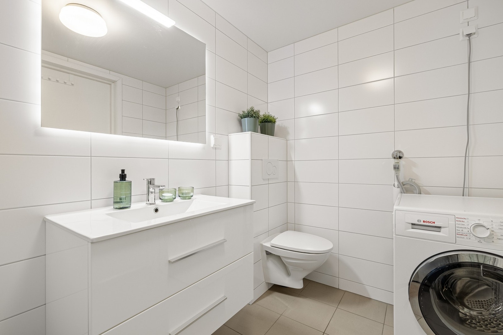 Badet er helfliset med underliggende varmekabler, og er innredet med vegghengt toalett, dusjhjørne med dører og baderomsmøblement med speil og belysning. Galleribilde