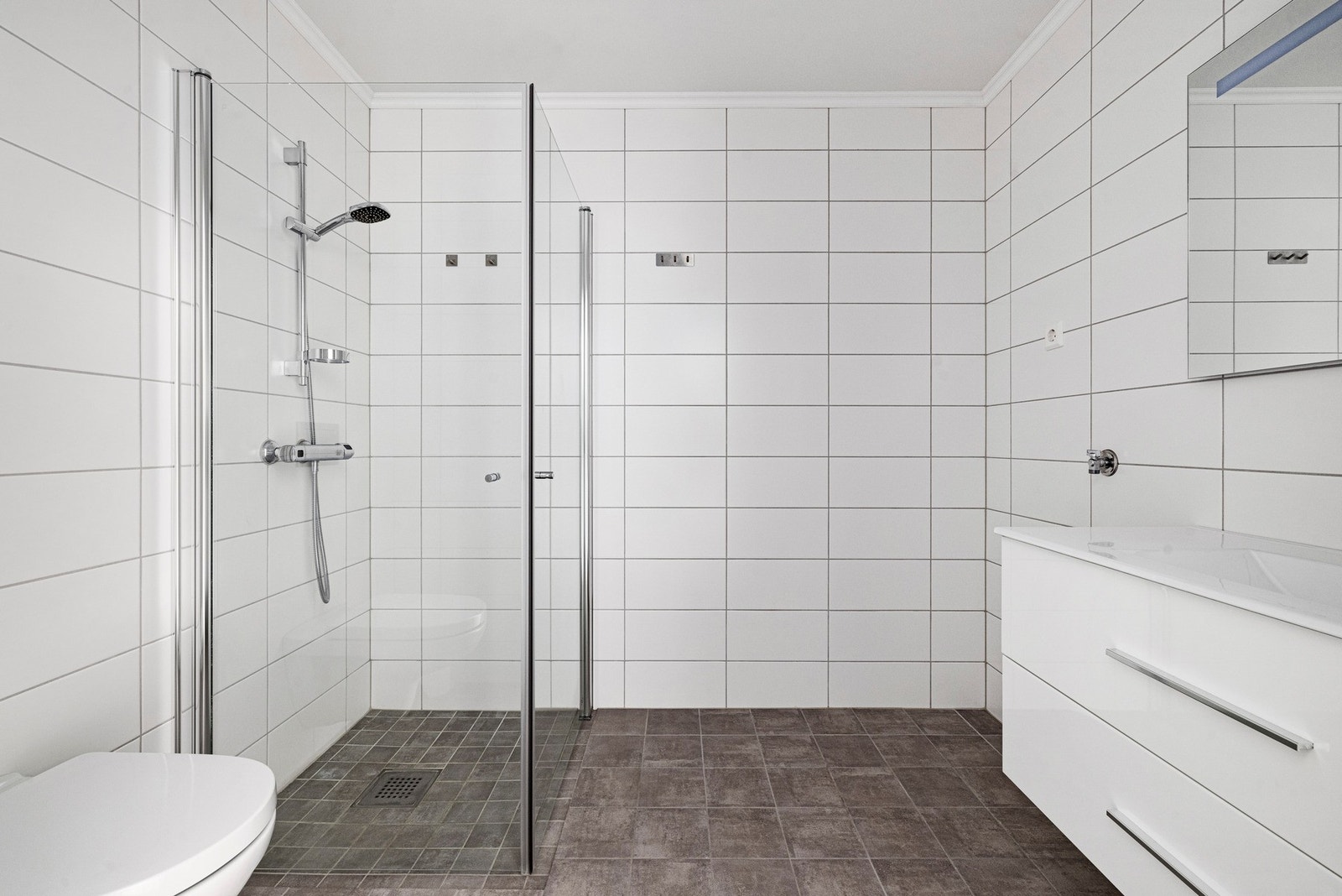 Flislagt bad med moderne uttrykk, dusj med glassvegger og praktisk oppbevaringsplass. Galleribilde