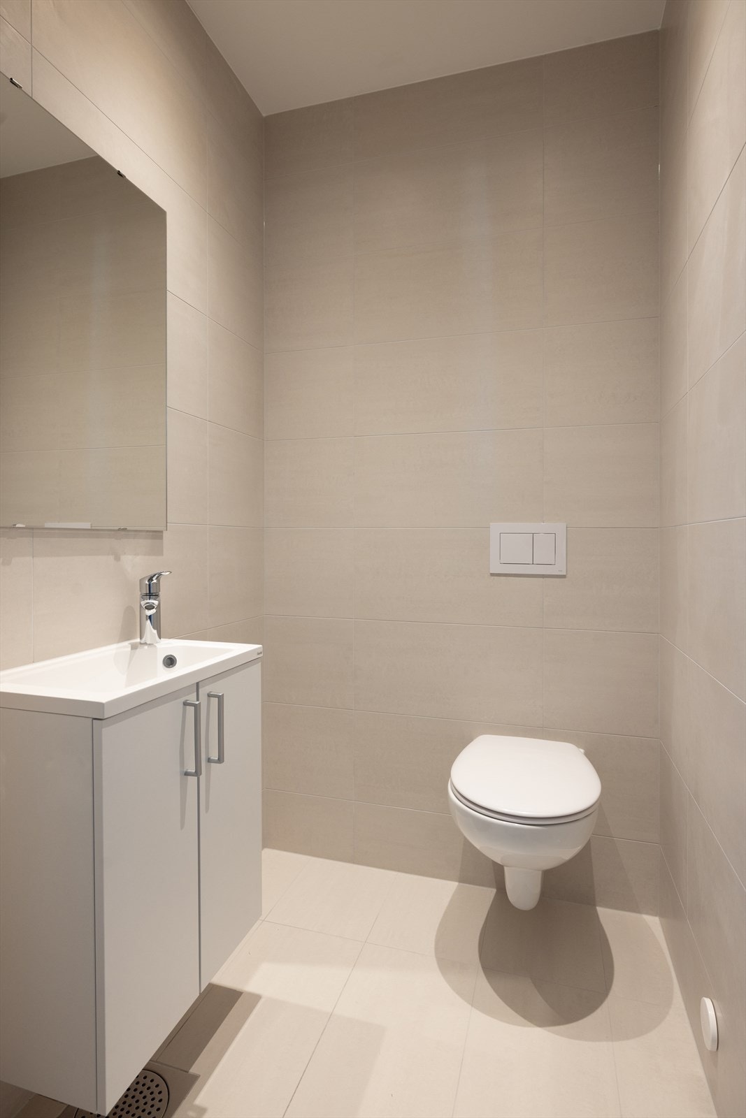 Pent, flislagt toalettrom med varmekabler i gulv, veggmontert toalett og servantskap med helstøpt servant, samt speil på vegg. Galleribilde