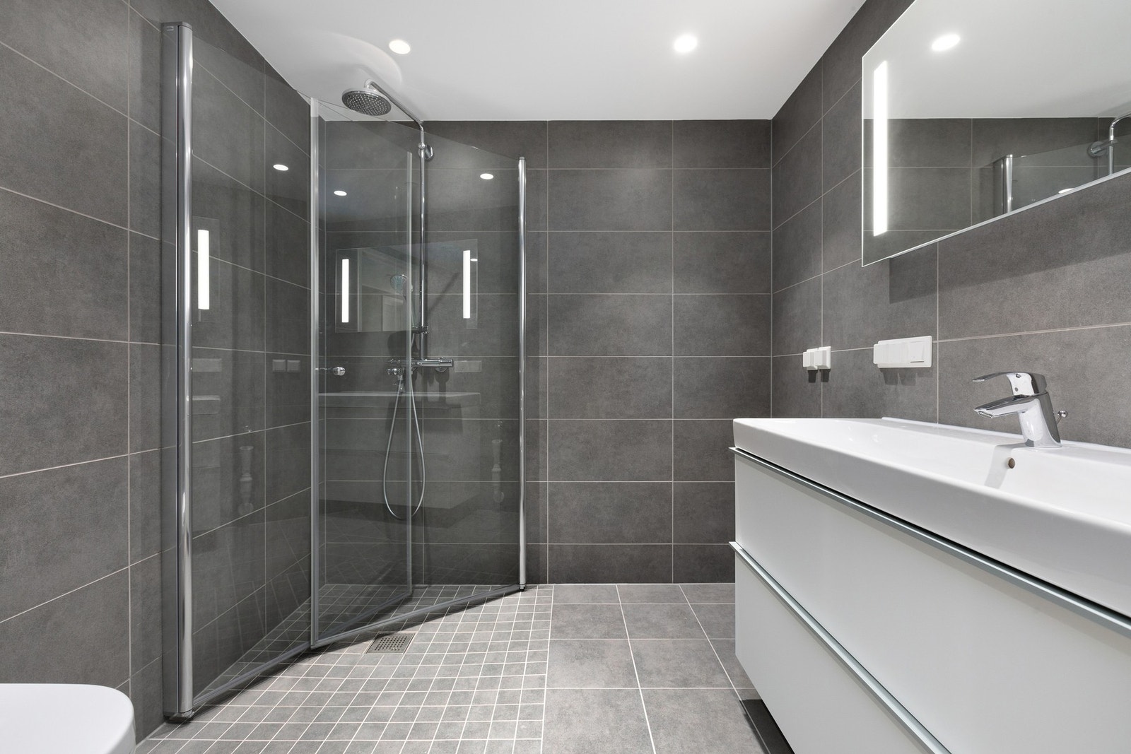 Badet fremstår stilrent og moderne med fliser på gulv og vegger, varmekabler i gulv og downlights i tak. Innredningen består av vegghengt baderomsinnredning med nedfelt servant, speil med belysning, vegghengt toalett samt dusjhjørne med innfellbare vegger Galleribilde