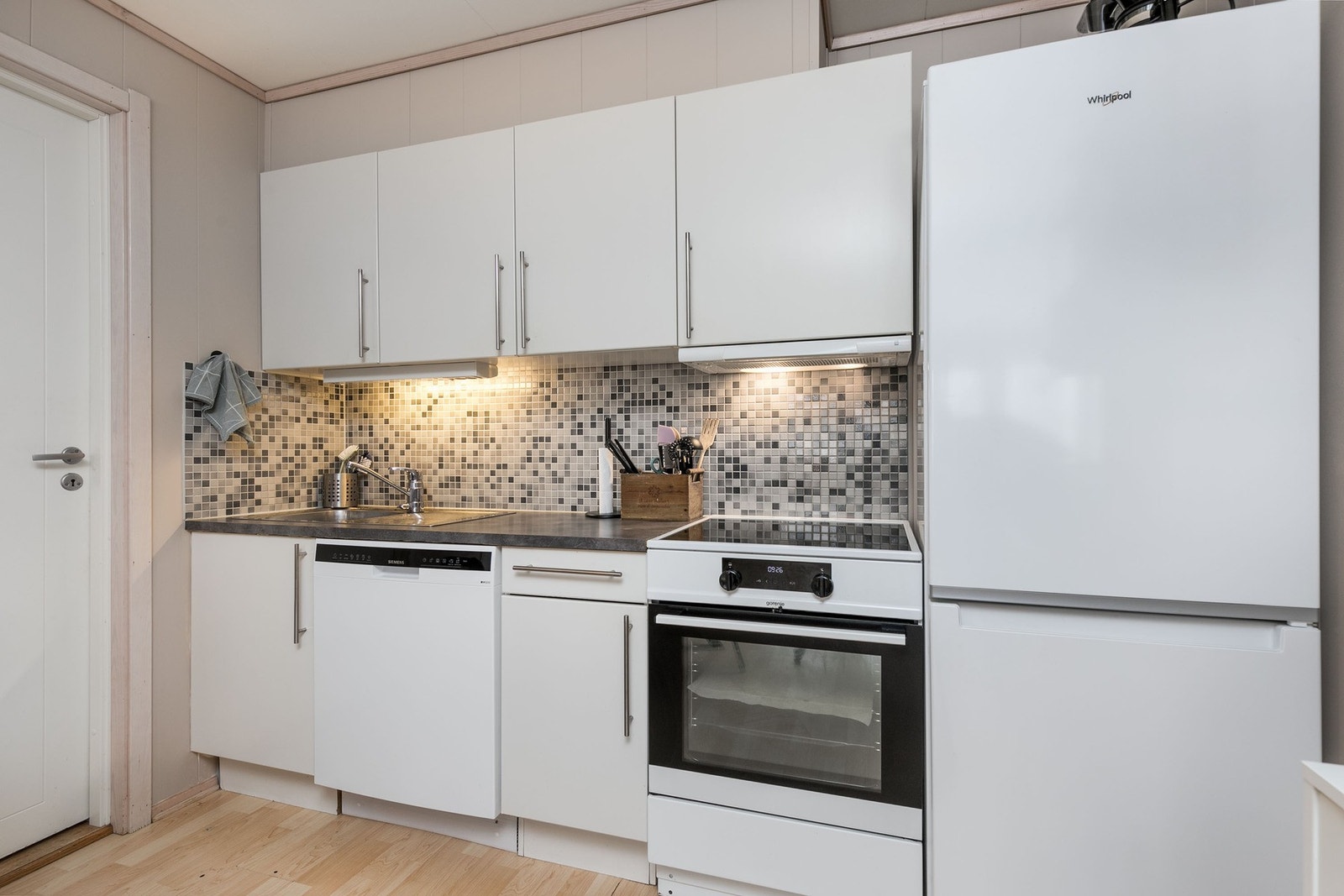 Kjøkkeninnredning med hvite fronter og håndtak. Frittstående komfyr, oppvaskmaskin og kjøleskap. Galleribilde