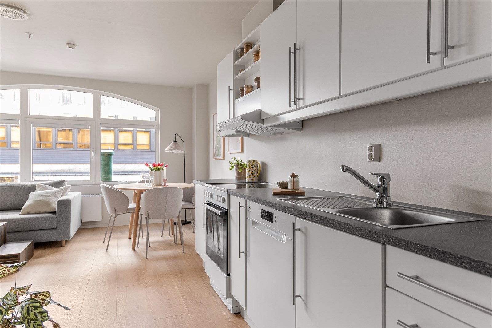 Kjøkkeninnredning med hvite fronter og laminat benkeplate med nedfelt stålvask. Kjøkkenet har ventilator og nisje for steikeovn, vaskemaskin og kjøleskap. Galleribilde