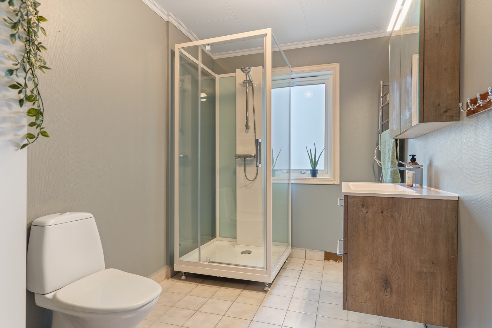 Det er fliser på gulv og malt strie på vegg.
Badet har dusjkabinett, seksjon med heldekkende vask og toalett. Galleribilde