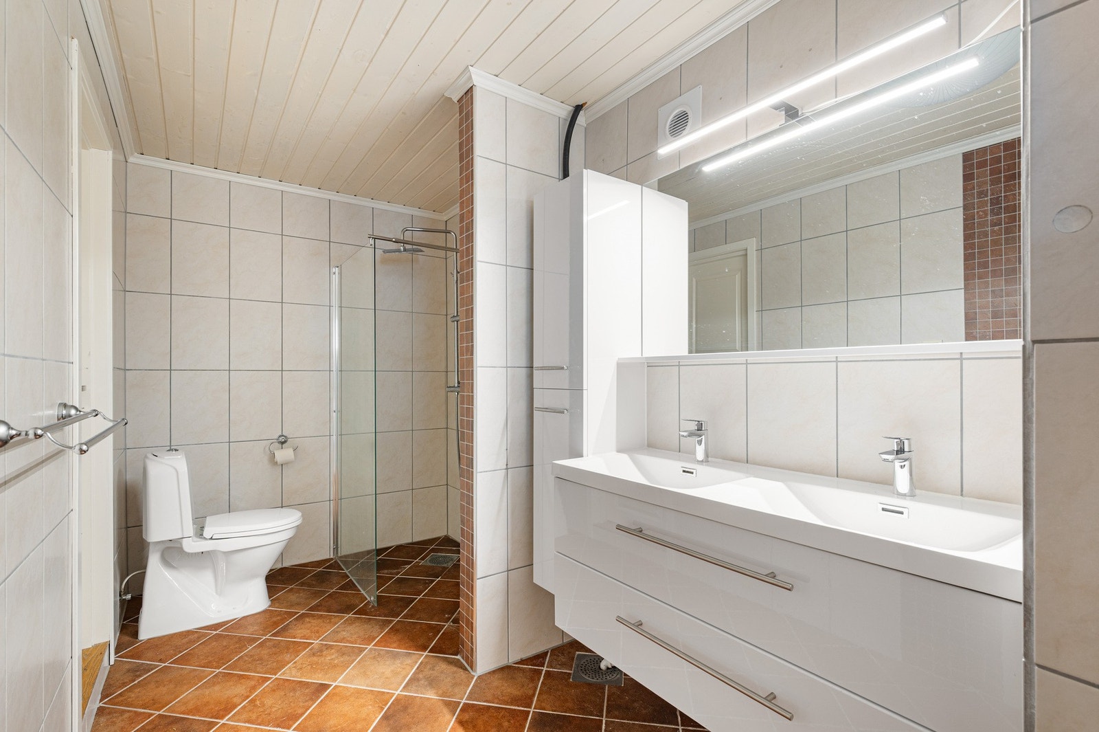 Romslig flislagt bad med baderomsinnredning, speil med lys, toalett og dusj. Galleribilde