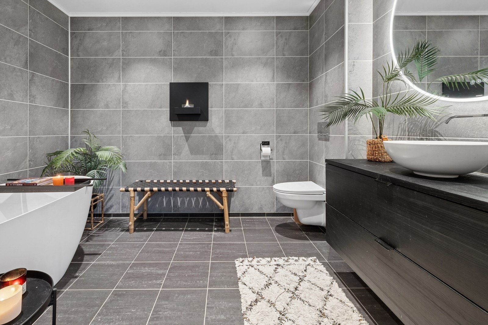 Badet i første etasje er innredet med badekar, vegghengt klosett og en sort vaskebenk med porselensservant. Badet har rundt speil med belysning og duggfjerning. Galleribilde