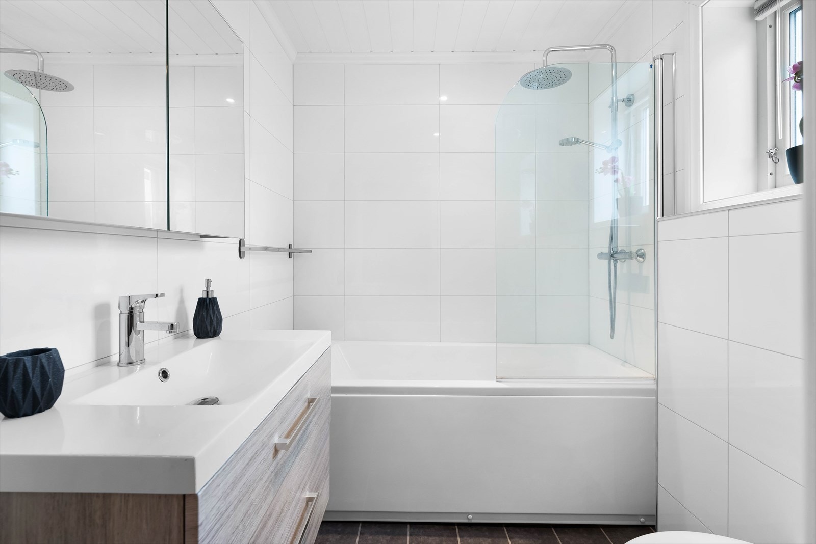 Badet er innredet med vegghengt toalett, badekar med dusj og glassdør og servant med skuffer. Galleribilde