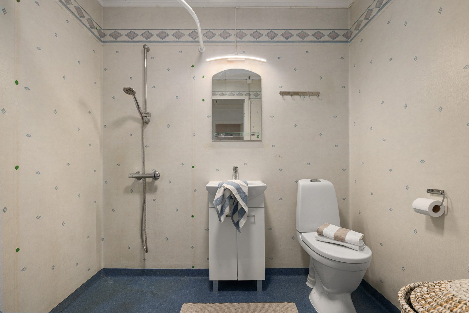 Badet er av eldre dato og har en enkel og funksjonell utforming. Rommet er innredet med servantinnredning med oppbevaringsplass, speil med belysning, gulvstående toalett og dusj med hånddusj montert på vegg. Galleribilde