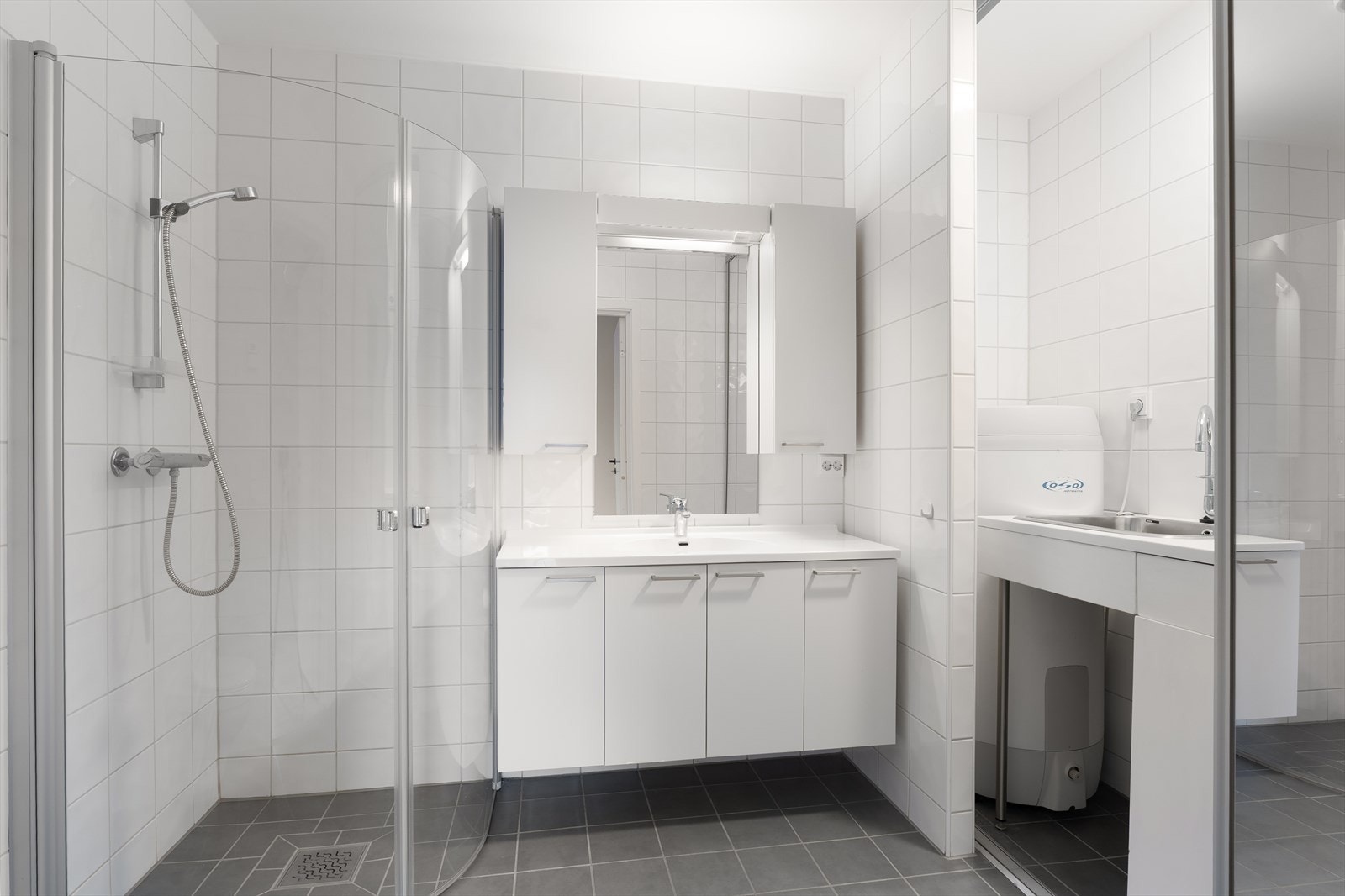 Leiligheten har et pent, helfliset bad som er innredet med hvit baderomsinnredning, dusjnisje med glassdører og vegghengt toalett Galleribilde