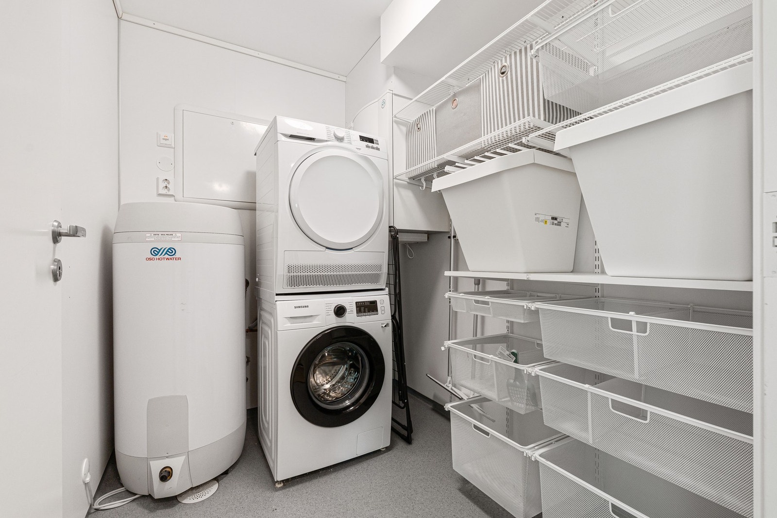 Praktisk vaskerom med opplegg for vaskemaskin, tørketrommel og oppbevaringshyller. Det er installert varmtvannsbereder. Galleribilde