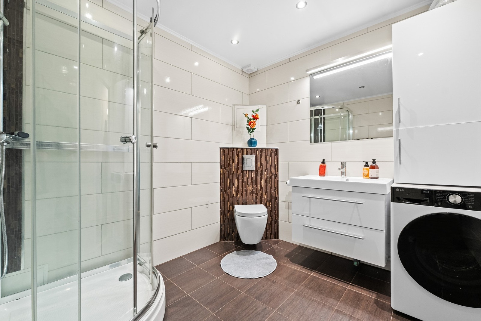 Innredningen består av servantmøbel med to skuffer, speil med toppmontert belysning og vegghengt toalett. Badet har et frittstående dusjkabinett, samt opplegg og plass for vaskemaskin og tørketrommel. Galleribilde