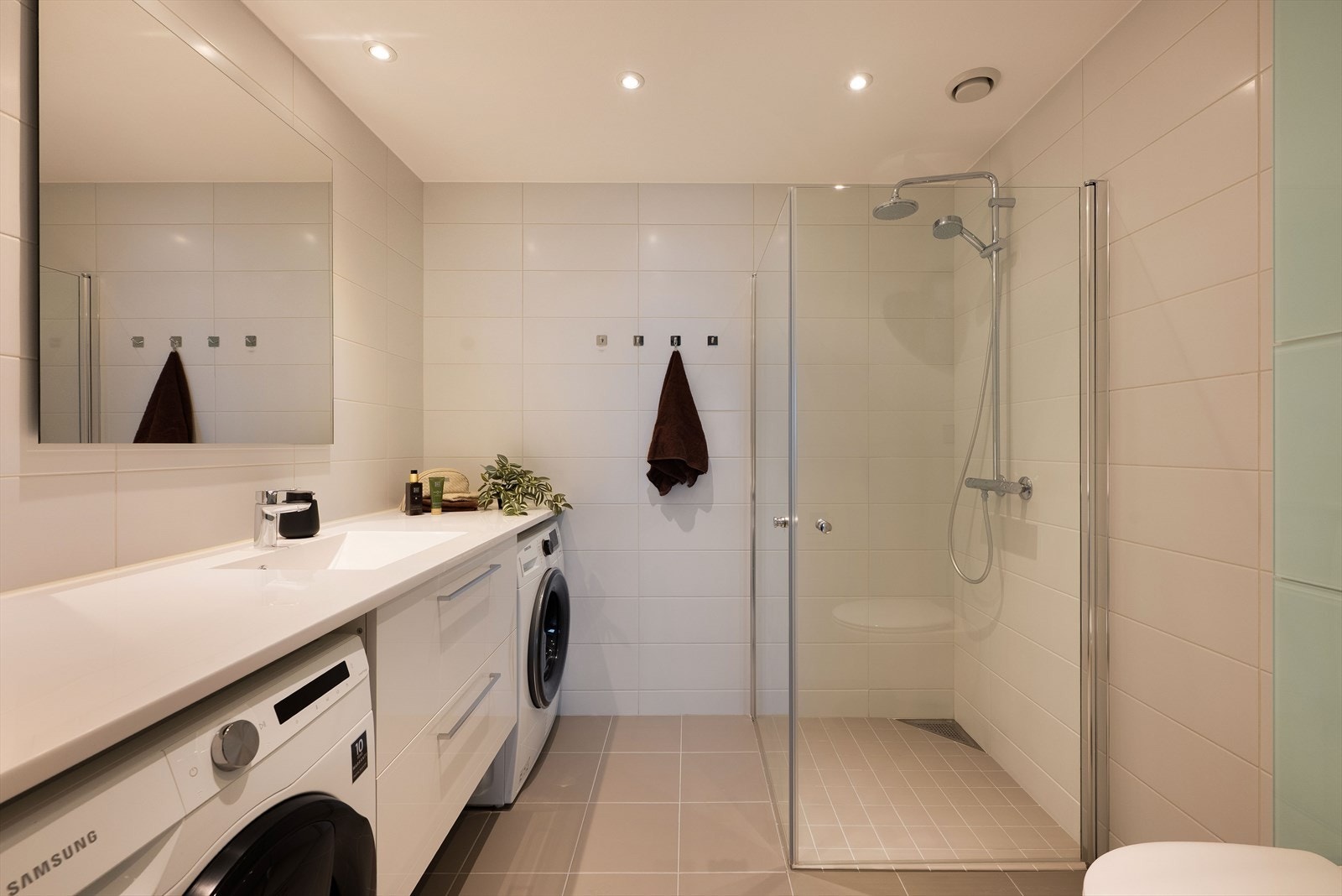 Pent baderom med tidløse fliser med vannbåren gulvvarme for ekstra komfort. Badet har et stilrent servantskap med glatte, folierte fronter og heldekkende servant. Over servant er det speil montert på vegg med lysarmatur. Galleribilde